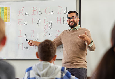 A teacher standing in front of a classroom of children while gesturing toward a whiteboard with the alphabet written on it