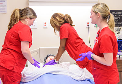 Three nursing students in red scrubs stand around a clinical dummy in a hospital setting