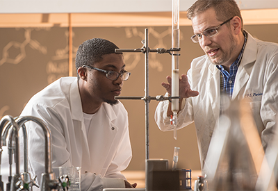 Two people in white labcoats in a lab environment observing a reaction in a test tube
